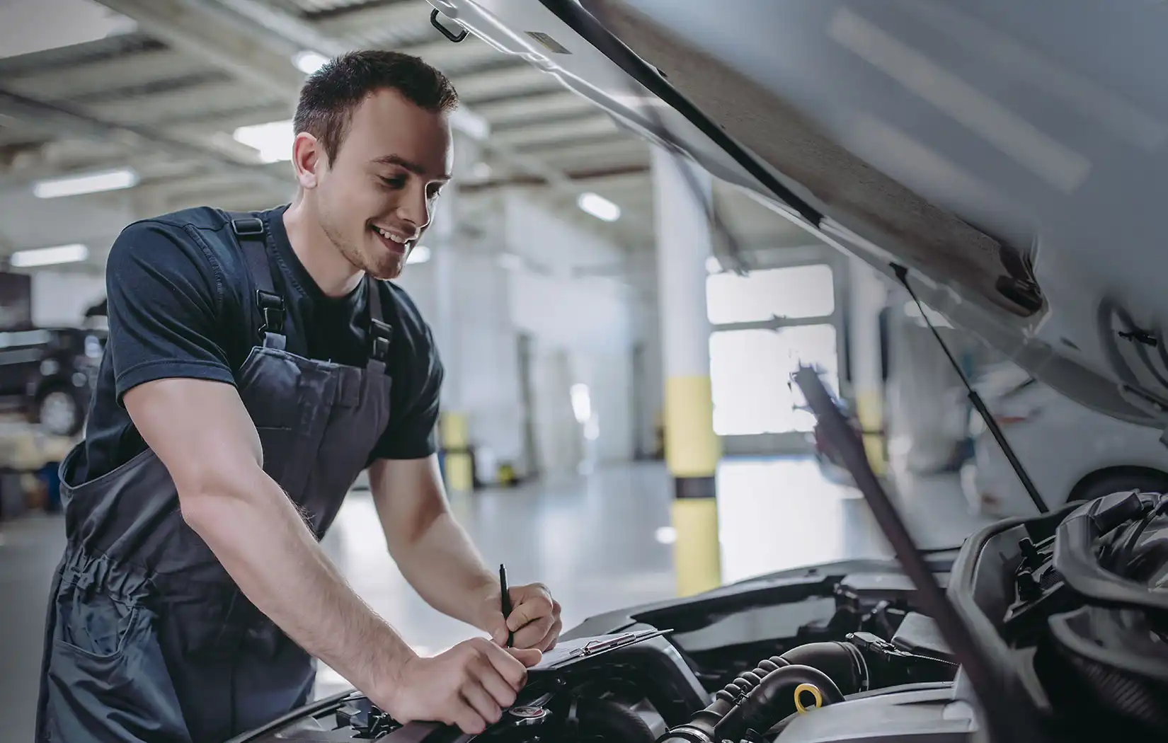 An auto technician writes on a clipboard next to a car with its hood raised.